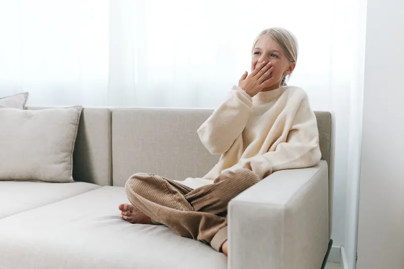 A young child sits on a light sofa, wearing a cream sweater and brown pants, covering her mouth with a hand in a candid, gentle moment of shy emotion.