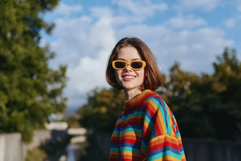 Woman smiling portrait outdoors in colorful striped sweater and yellow sunglasses, happy young adult enjoying sunny day, candid fashion lifestyle and cheerful expression.