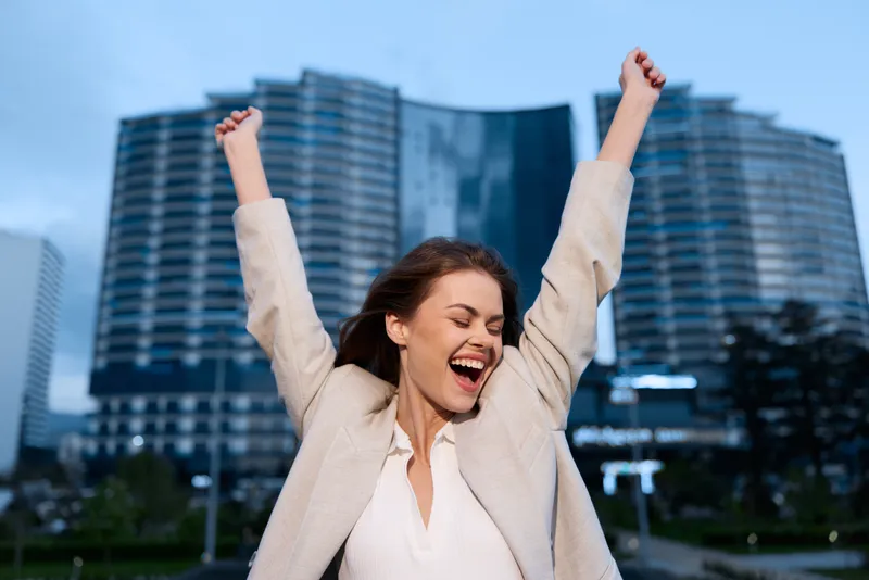 Excited woman celebrating financial success outdoors in modern urban business district, confident female with arms raised in beige jacket and white blouse