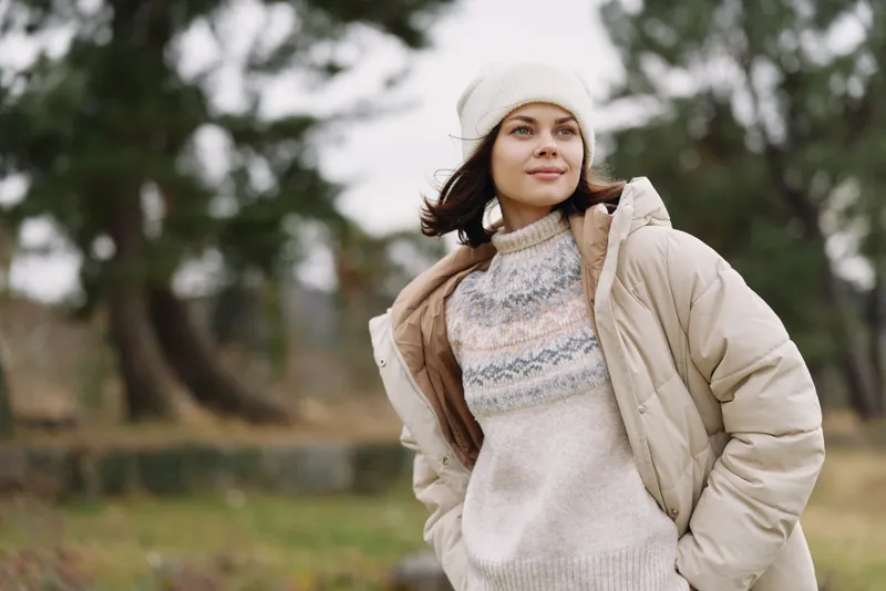 Woman in a beige coat and knit sweater stands outdoors in a park, wearing a beanie. Calm scene conveys cozy winter fashion with soft light and relaxed attitude.