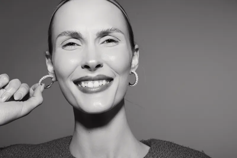 Smiling white woman with fair skin wears hoop earrings and knitted sweater, looking joyful in studio portrait with neutral background.