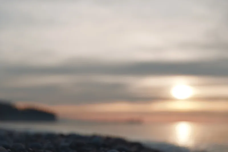 Sunset over a tranquil beach and calm sea with a soft glow on the horizon; blurred shoreline, gentle waves and muted colors create a peaceful coastal mood.