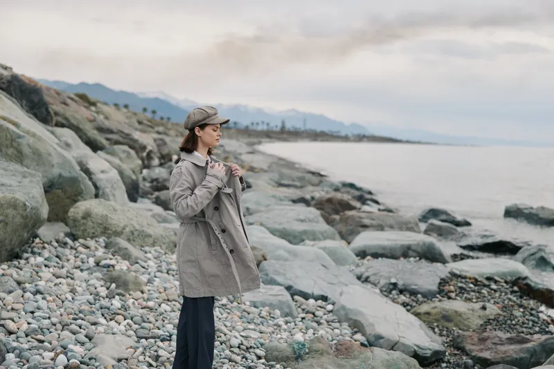 Young woman in a plaid coat and hat stands on a rocky beach looking at the sea. Cloudy sky and mountains create a calm and contemplative mood by the shore.