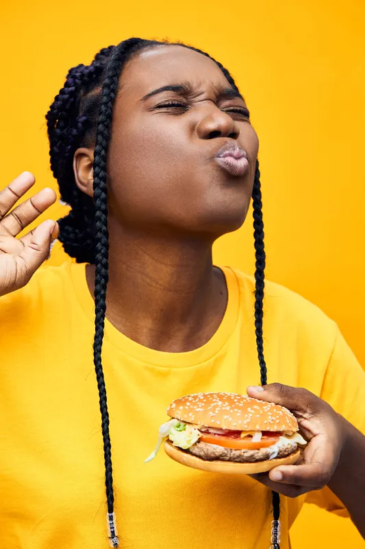 Joyful young woman enjoying a delicious burger against a bright yellow background, wearing a casual yellow shirt, conveying happiness and satisfaction