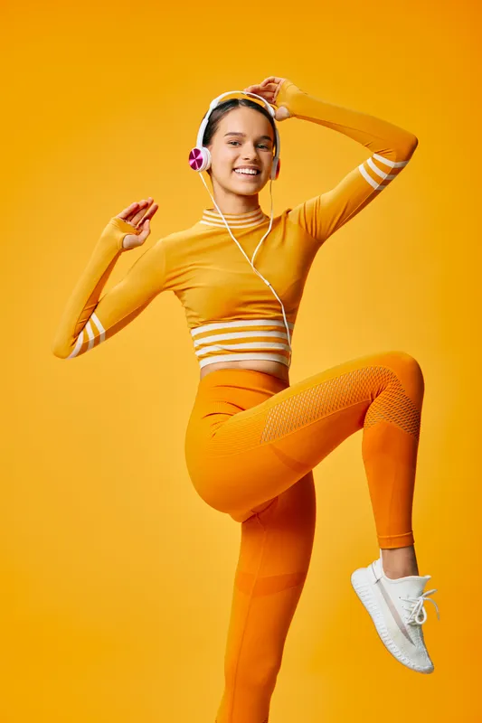 Energetic woman fitness dancer in orange athletic wear with headphones, leaping in a dynamic pose against a vivid studio backdrop, showcasing movement, balance and athletic style.