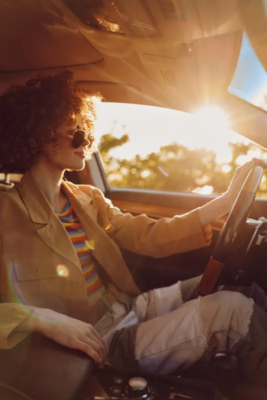Cheerful woman driving a car at sunset, casual chic, sunlit moment, relaxed posture, warm tones, retro film color vibe, outdoors, vibrant colors, lifestyle portrait, natural setting