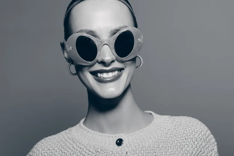 Smiling young caucasian woman with pale skin wearing large round sunglasses and hoop earrings. She wears a knitted sweater on grey background, fashion and style concept.
