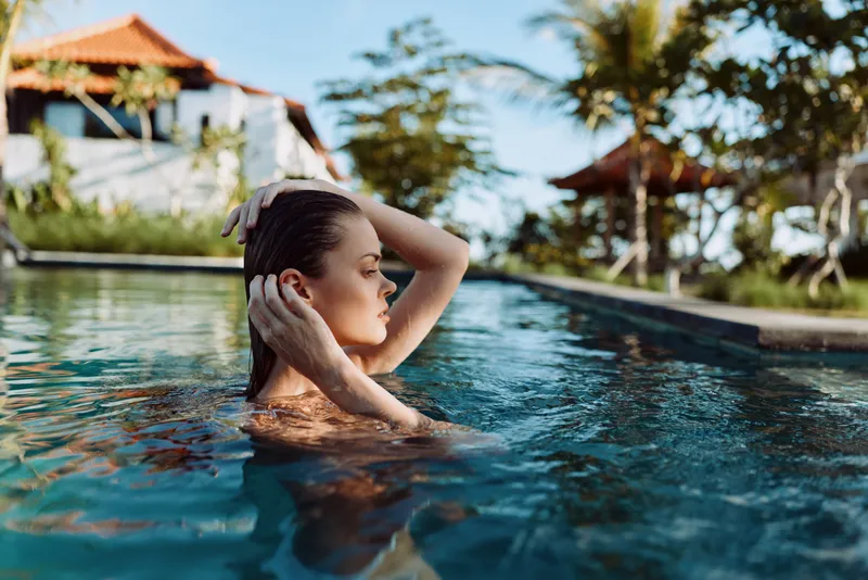 Young woman with long hair relaxes in a swimming pool at a tropical resort on a sunny day, enjoying a peaceful moment with her eyes closed and hand touching her head.