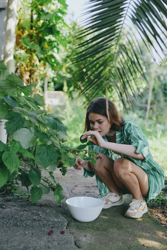Young woman crouches in a lush garden, gathering fresh produce into a white bowl amid vibrant leaves and tropical plants.