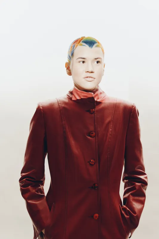 Young woman with colorful short hair wearing a red leather jacket poses confidently against a minimalist light background in a modern fashion style.