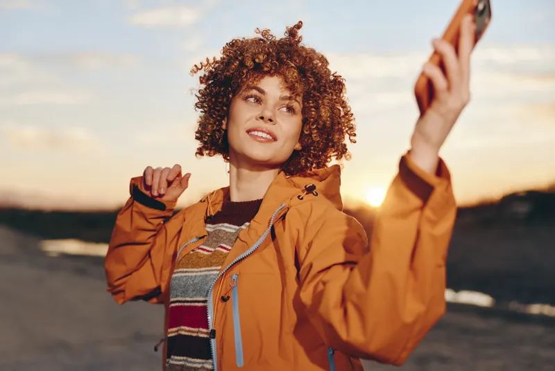 Woman with curly hair in orange jacket outdoors at sunset, holding phone for selfie, warm golden light and soft wind, lifestyle travel portrait, casual smile