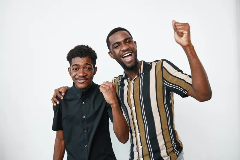 Two African American men with dark skin smiling and celebrating together in casual clothes on white background. Friendship, happiness and positive emotions in studio portrait.