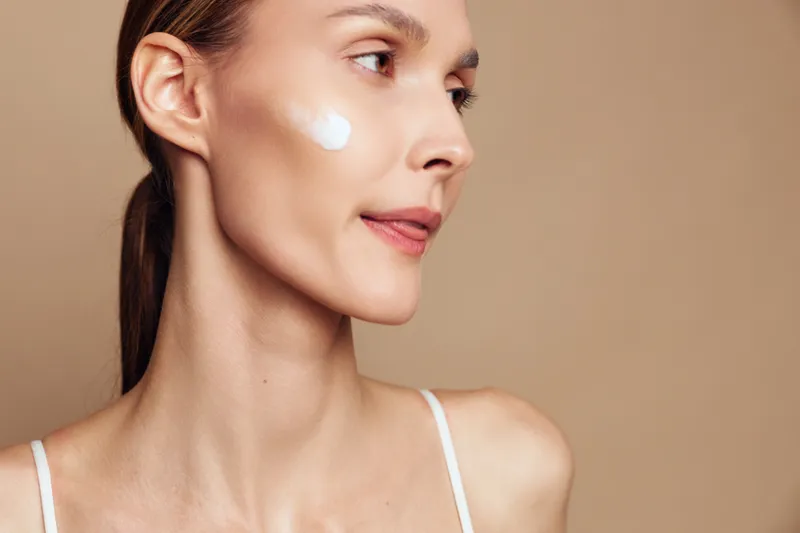 Skincare woman beauty face close up shows white cream on cheek, calm expression with side gaze, studio lighting, beige background, skincare treatment concept for hydration and self care now