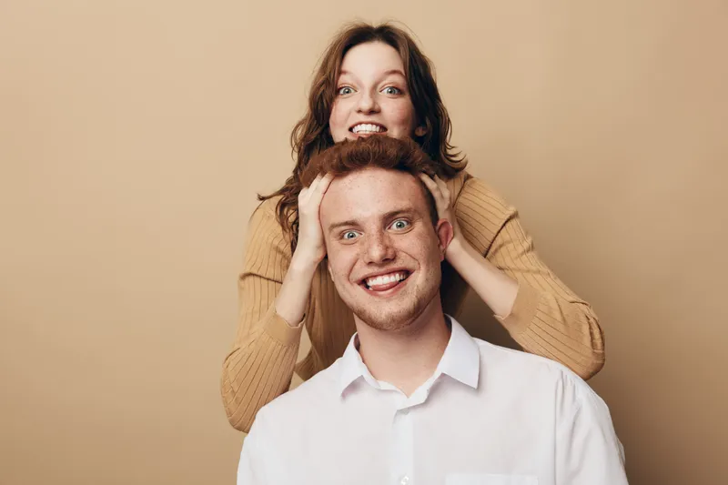 Happy young man and woman playfully pose together on beige background. The woman stands behind the man with hands on his head, both smiling joyfully. Concept of friendship and fun.