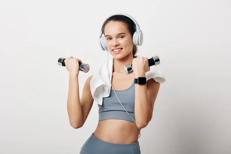 Young woman in fitness outfit lifting light dumbbells, wearing headphones and towel over shoulders, smiling confidently in bright studio, sport workout concept.