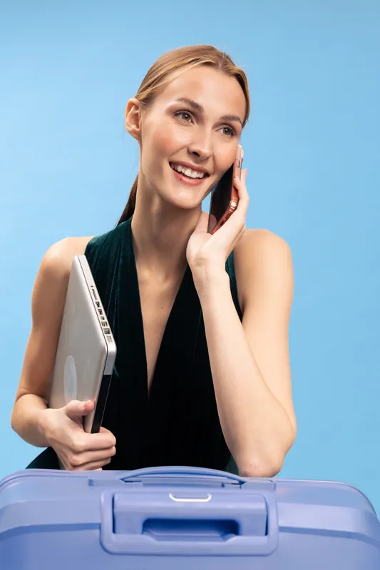 Smiling young woman holding laptop and talking on phone with blue suitcase in front. Concept for travel, business trip, remote work, and communication on light blue background.