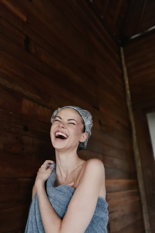 Laughing woman wrapped in towel with shower cap stands against wooden wall indoors, expressing happiness and freshness after shower, natural beauty, relaxed mood.
