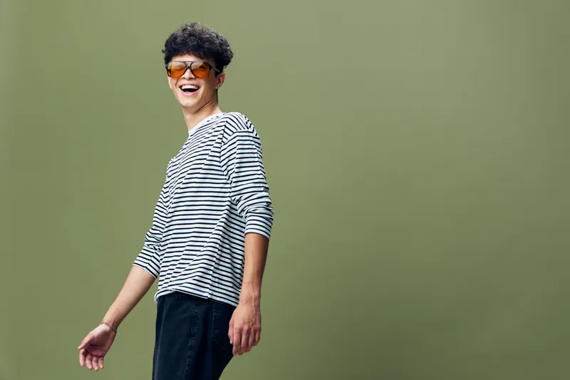 smiling man wearing sunglasses and striped shirt stands against green background with casual pose and joyful expression, young adult in modern style