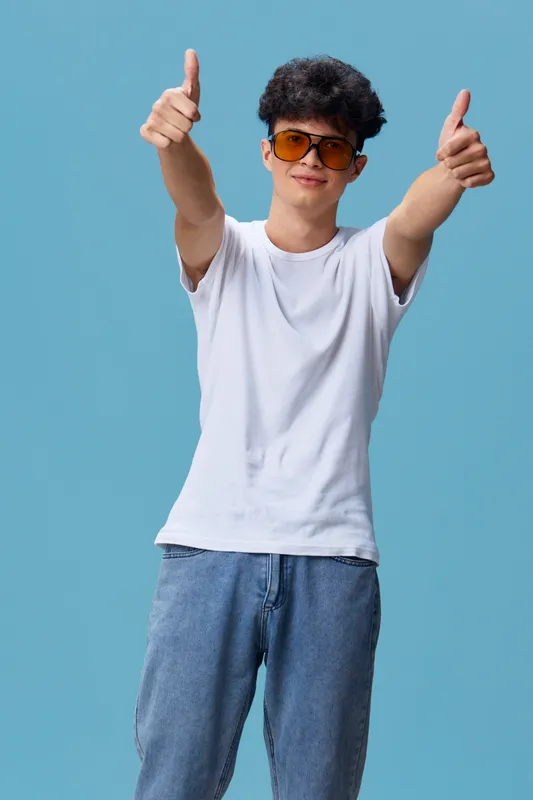 Young man wearing orange sunglasses and casual white t-shirt shows thumbs up gesture with both hands, smiling confidently against a solid blue background.