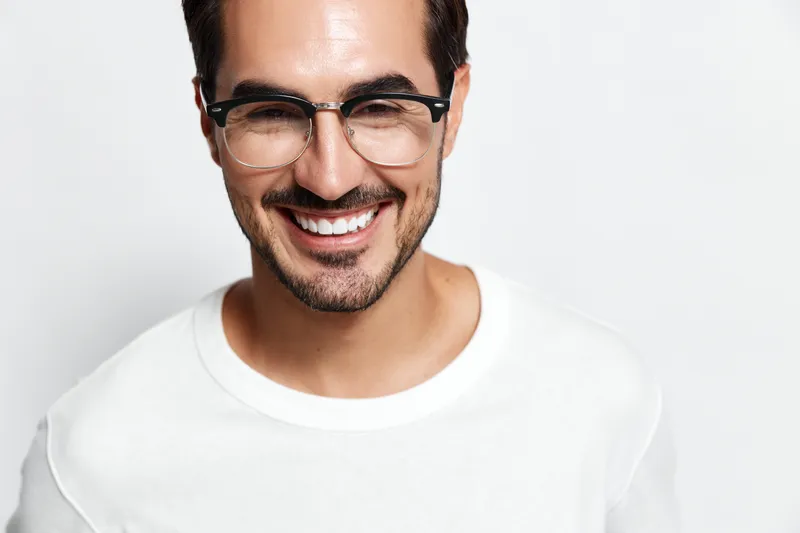 Happy cheerful smiling young man wearing glasses, looking at the camera with a bright smile, dressed in a casual white t-shirt against a solid light background.