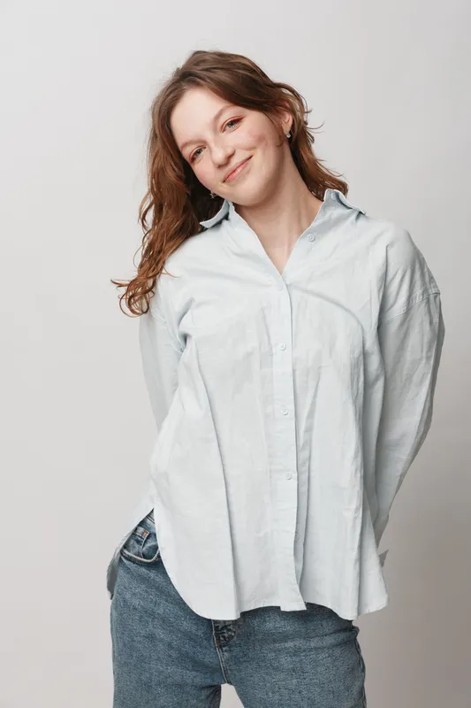 Smiling young woman with long brown hair wearing a white shirt and blue jeans stands against a light background. Casual style, natural beauty, and relaxed confident mood concept.
