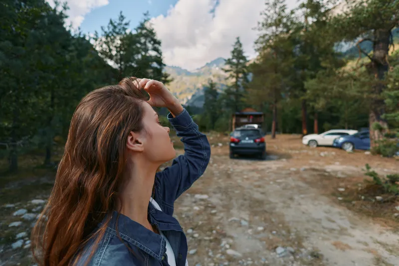 A woman wearing a denim jacket stands in a forest parking area, looking up as she shields her eyes from the sun, enjoying the outdoors and a bright sky.