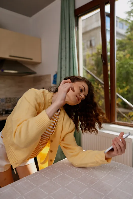 Smiling woman leaning on a kitchen table in a yellow cardigan, checking her phone in a bright home setting, presenting casual lifestyle, warmth and everyday comfort.