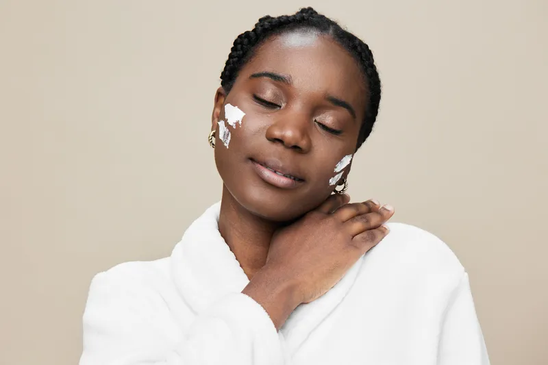 Calm woman with dark skin in a white robe applying skincare on her face, showcasing a healthy lifestyle and self care routine in a minimalistic beige background