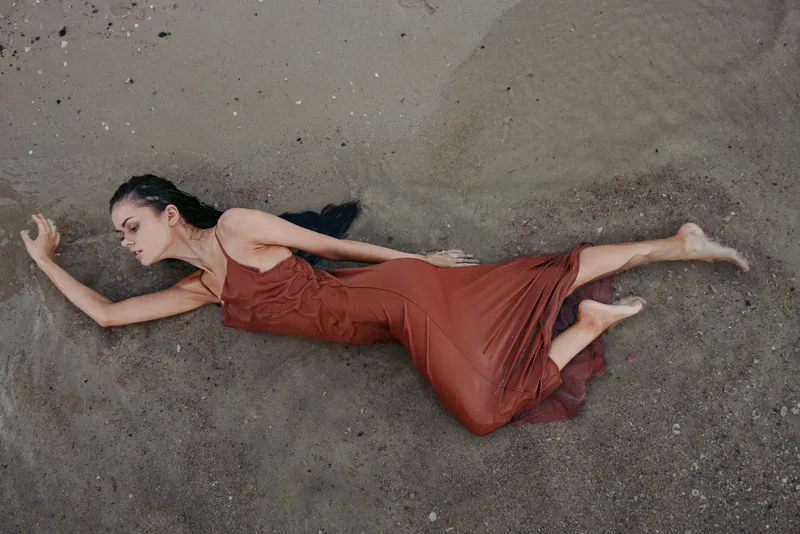 Woman lying on sandy beach wearing red dress with closed eyes, expressing calm and relaxation in natural outdoor setting.