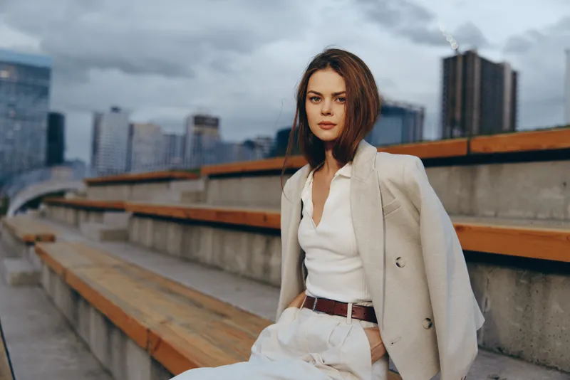 Confident woman in business attire sitting outdoors, showcasing professionalism against an urban skyline backdrop with soft colors and a modern style.