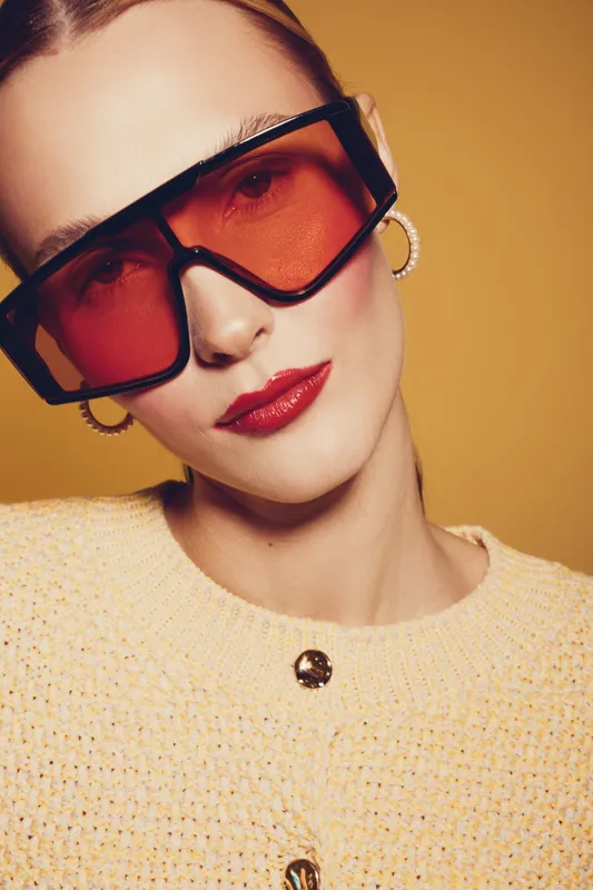 Close-up portrait of a stylish woman wearing oversized red sunglasses and hoop earrings. She wears a textured yellow sweater against a warm orange background. Fashion and accessories concept.