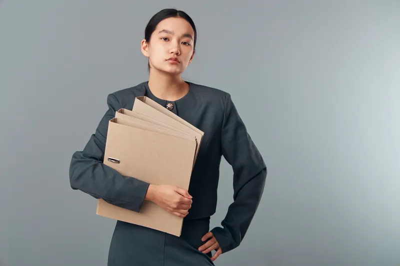 Young woman in dark business attire holding multiple cardboard folders, standing on neutral background with serious expression, symbolizing office work and organization.