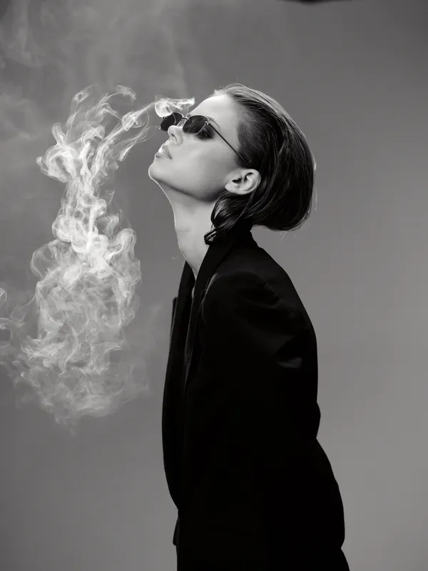 Black and white portrait of a stylish woman with sunglasses exhaling smoke, dressed in a dark jacket with a minimalist background.
