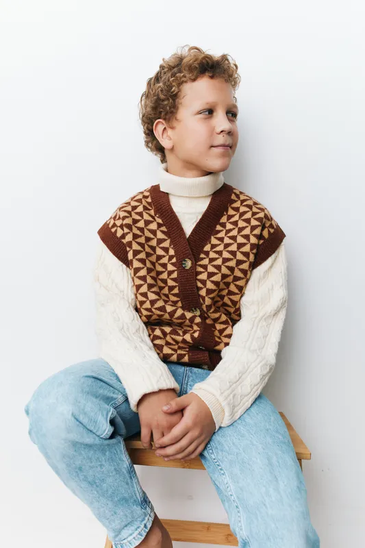 Young boy sits on a wooden bench wearing a cozy knit vest over a cream turtleneck, looking off to the side with a calm, thoughtful expression, framed by soft studio light.
