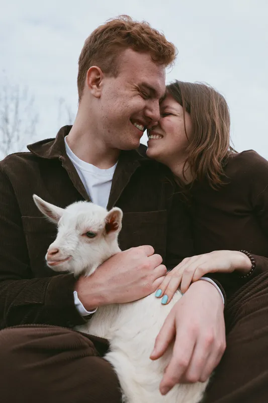 Casual couple outdoors with a white goat kid, sharing affection and warm smiles, capturing a tender lifestyle moment, rustic setting, cozy atmosphere, and genuine happiness.