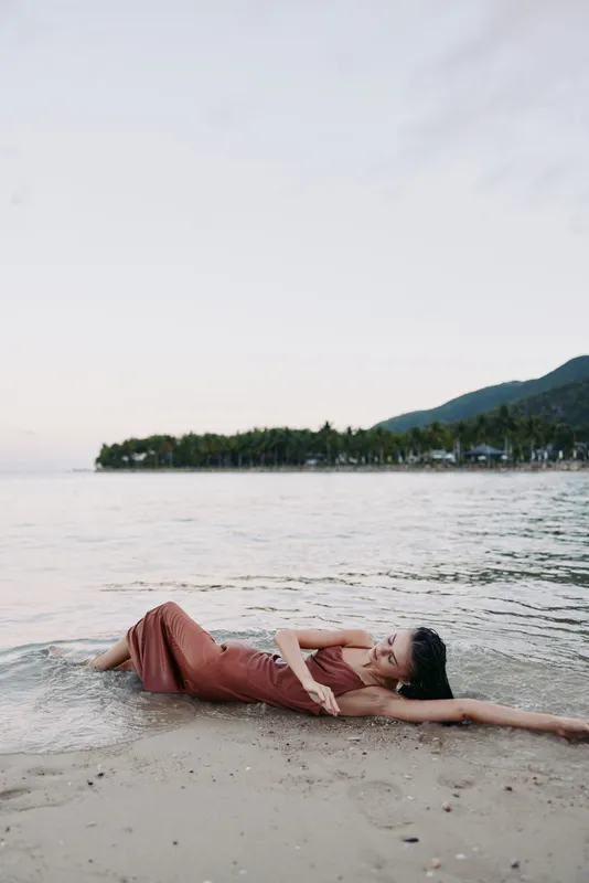 A person reclines on a sandy shore beside a calm sea, wearing a rust red dress, with arm extended, creating a serene, relaxed focal point on the beach.