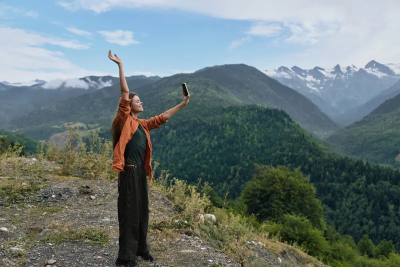 Woman stands on a rocky overlook taking a selfie with a dramatic mountain landscape, raised arms, clear blue sky and distant peaks behind ahead