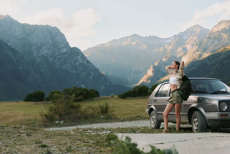 A young woman traveler stands beside a small car on a mountain road, arms raised in triumph as towering peaks loom in the distance, capturing a moment of adventure.
