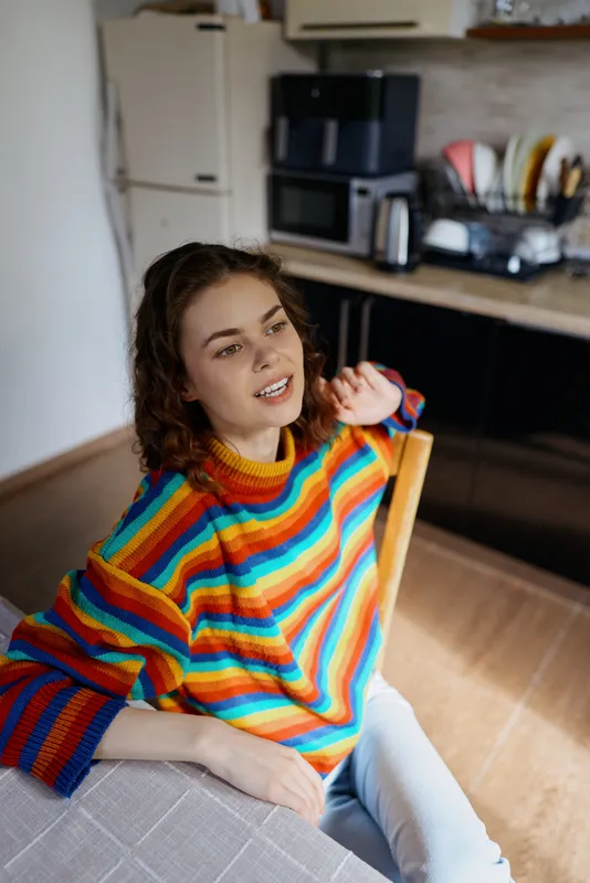 Cheerful woman indoors wearing a colorful striped sweater, relaxing at home, sunlit natural light, warm kitchen backdrop, candid smile, casual pose, cozy atmosphere, soft colors, everyday lifestyle.