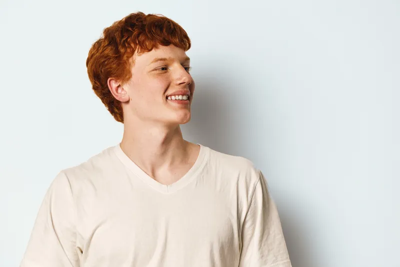 Young man with red hair and freckles smiling and looking to side in casual light t-shirt on white background. Portrait of happy male person with natural expression and short hairstyle.