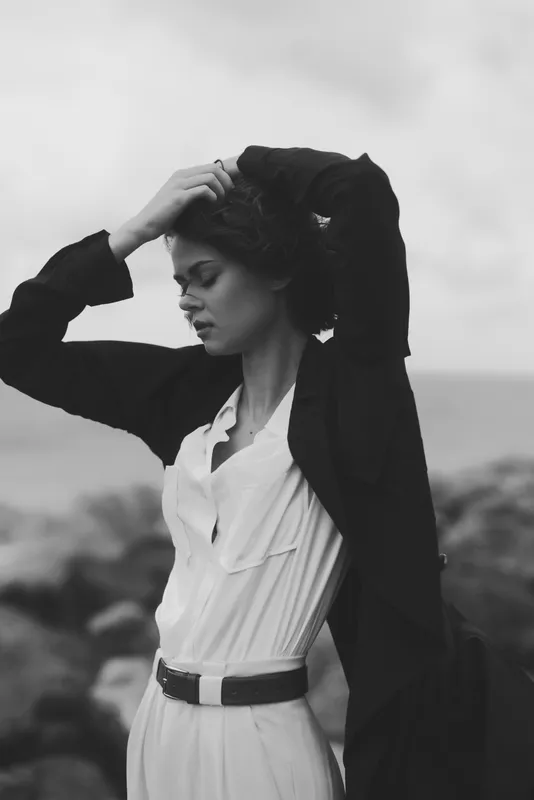 Woman in a flowing white dress and dark coat stands outdoors, hands in hair, wind swept, moody monochrome fashion moment of confidence and elegance, timeless poise