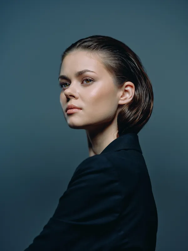 Portrait of a young woman with short brown hair wearing dark clothing against a simple blue-gray background, looking confident and thoughtful.