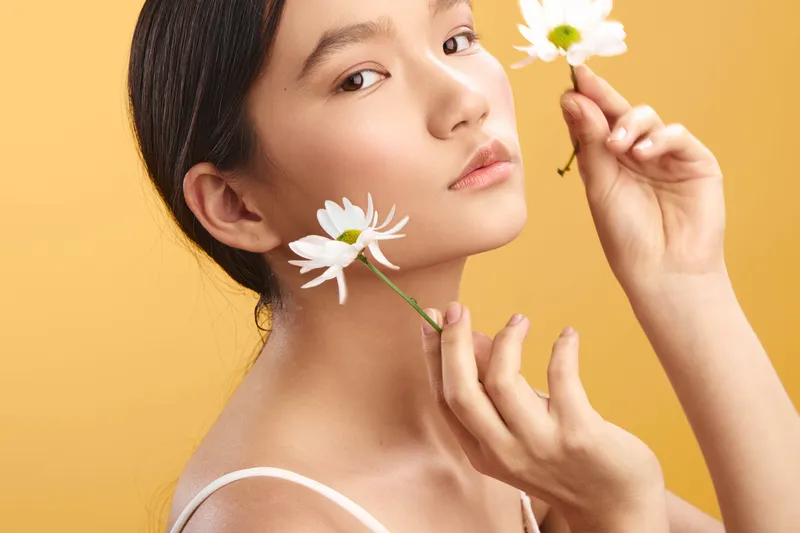 asian woman gazes softly at the camera, holding white flowers near her face in a studio portrait with a warm yellow background, conveying delicate beauty and calm, inviting mood.