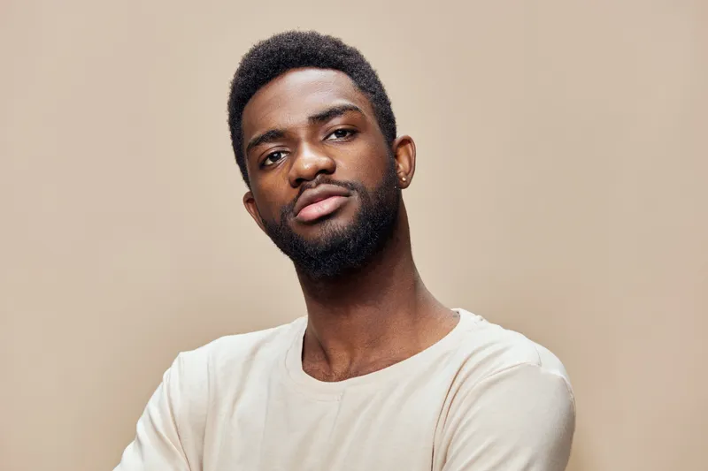 Confident young African man with stylish hair wearing a cream shirt, posing against a neutral beige background, showcasing self assurance and modern style