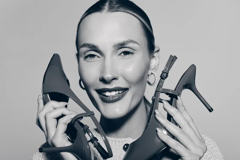 Smiling white woman with light skin holds two high heel shoes near face, wearing hoop earrings and knitted sweater. Concept of fashion, style, and footwear.
