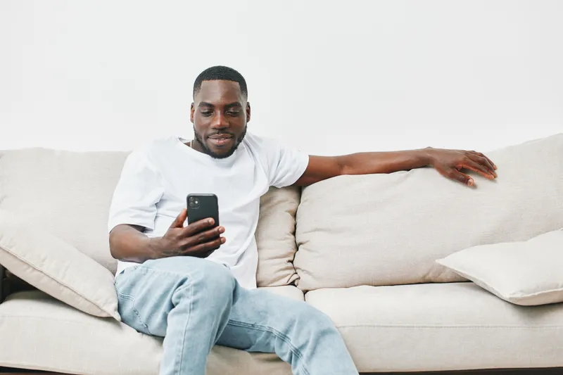 Relaxed African man using smartphone on a light colored couch, expressing joy and engagement, perfect for lifestyle and technology themes