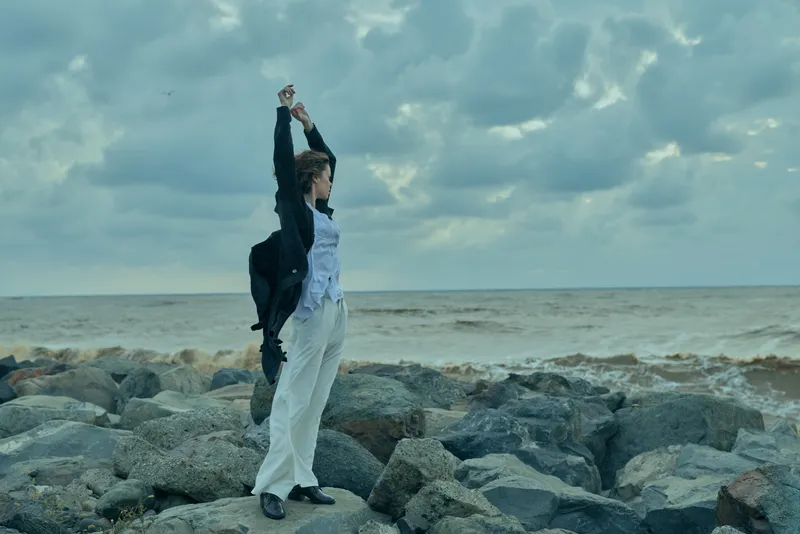 Woman standing on a rocky shore with arms raised, facing the sea, wearing light trousers and a jacket, conveying freedom, resilience and outdoor exploration by the ocean.