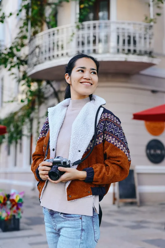 Photography camera street style outdoor shoot featuring a smiling woman in a warm brown jacket holding a camera in a lively urban street with cafes flowers and city vibe