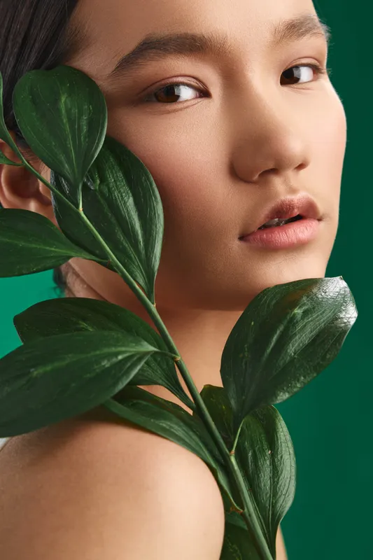 asian woman with lush leaves framing her face in a closeup portrait, soft lighting highlights smooth skin and calm expression, natural beauty exudes freshness and elegance.