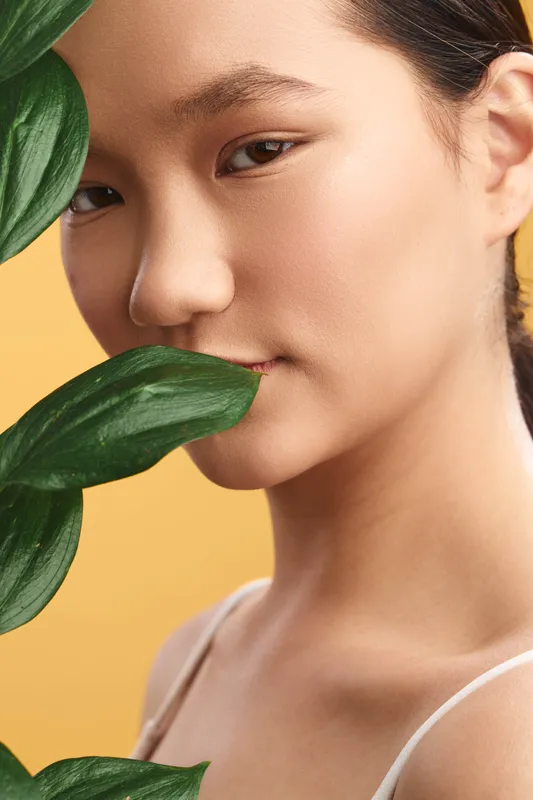 Asian woman with smooth skin appears in a close up portrait, framed by lush green leaves and a warm yellow backdrop, conveying calm elegance and natural beauty.